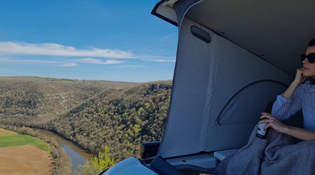 Photo du Van avec le toit ouvert sur un spot pour admirer la vue - une femme est assise avec lunette de soleil et boisson à la main elle regarde la vue - ma petite vadrouille